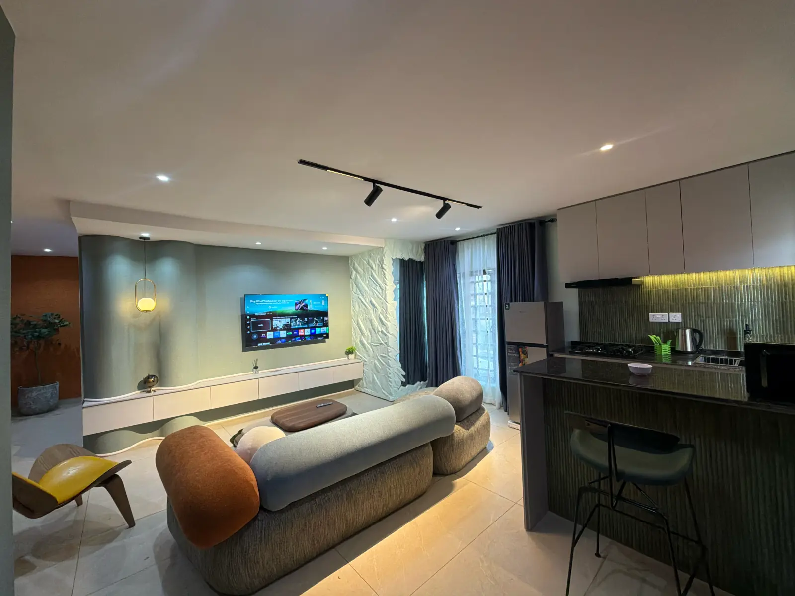The living room and kitchen: curved grey sofas, a yellow lounge chair, large portraits and a green-tiled kitchen beyond