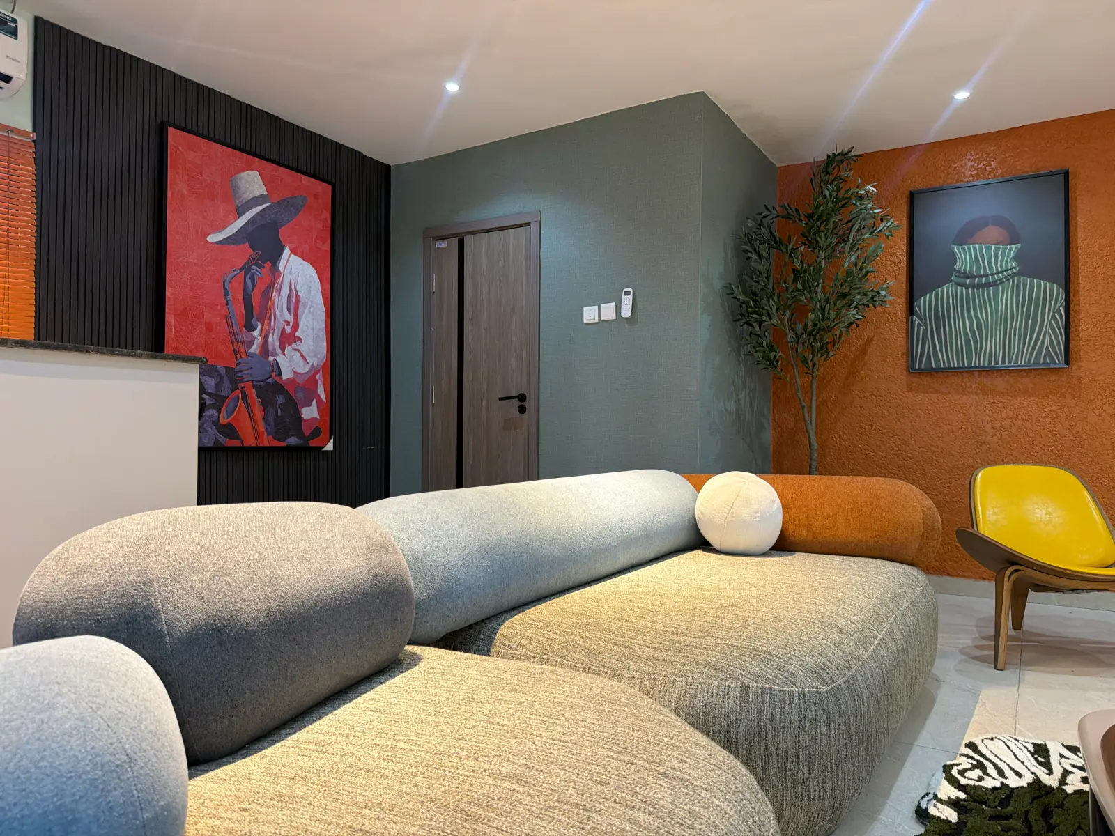 Living room with a grey curved sofa, orange bolster, yellow lounge chair and two large framed portraits