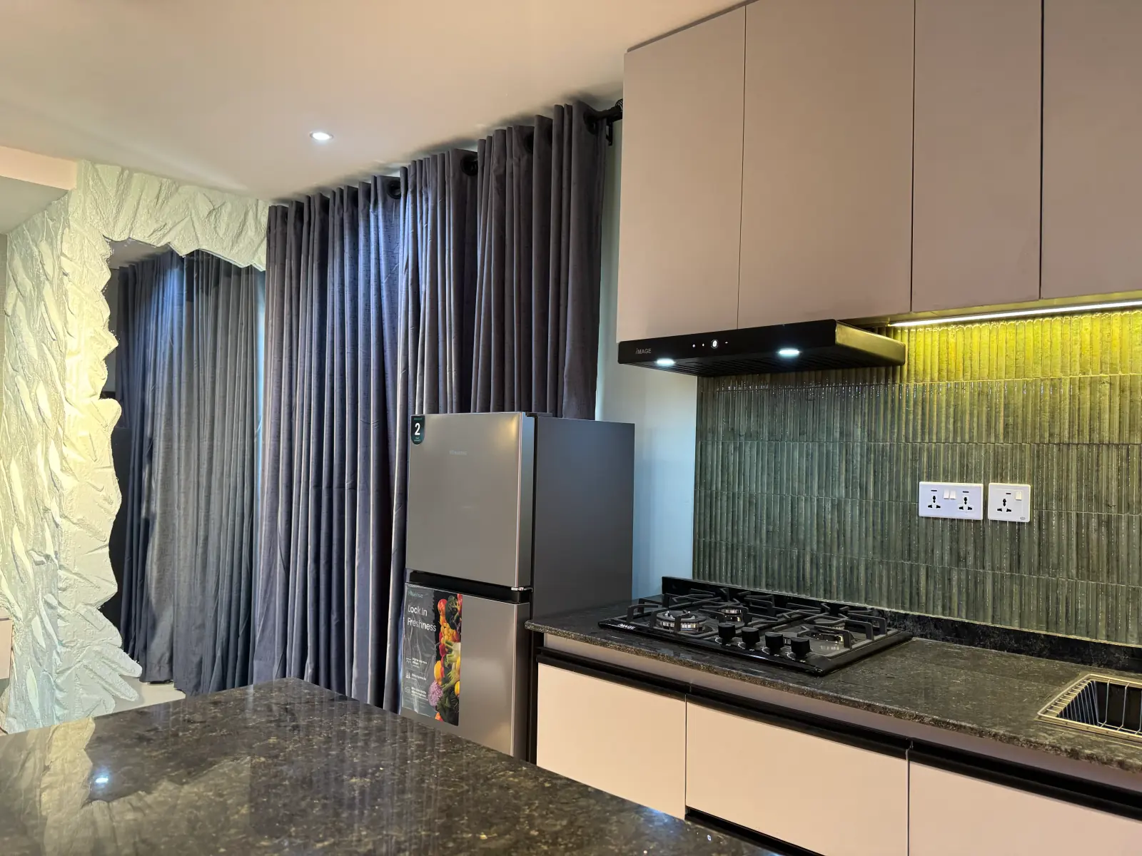Kitchen with a black gas hob, grey-green tiled splashback, steel fridge and grey curtains