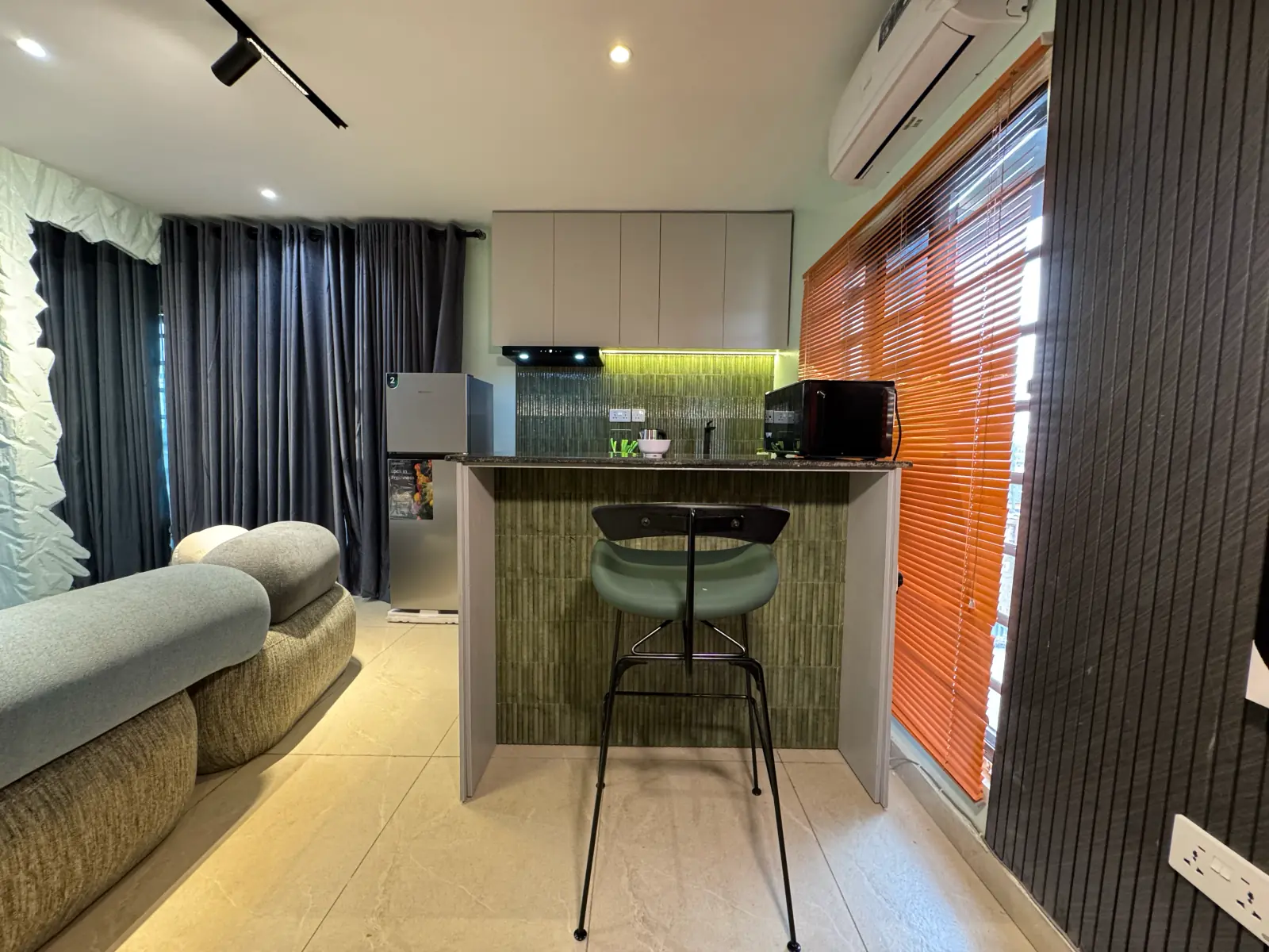 The breakfast bar and kitchen seen from the living room, with grey curtains and an orange blind